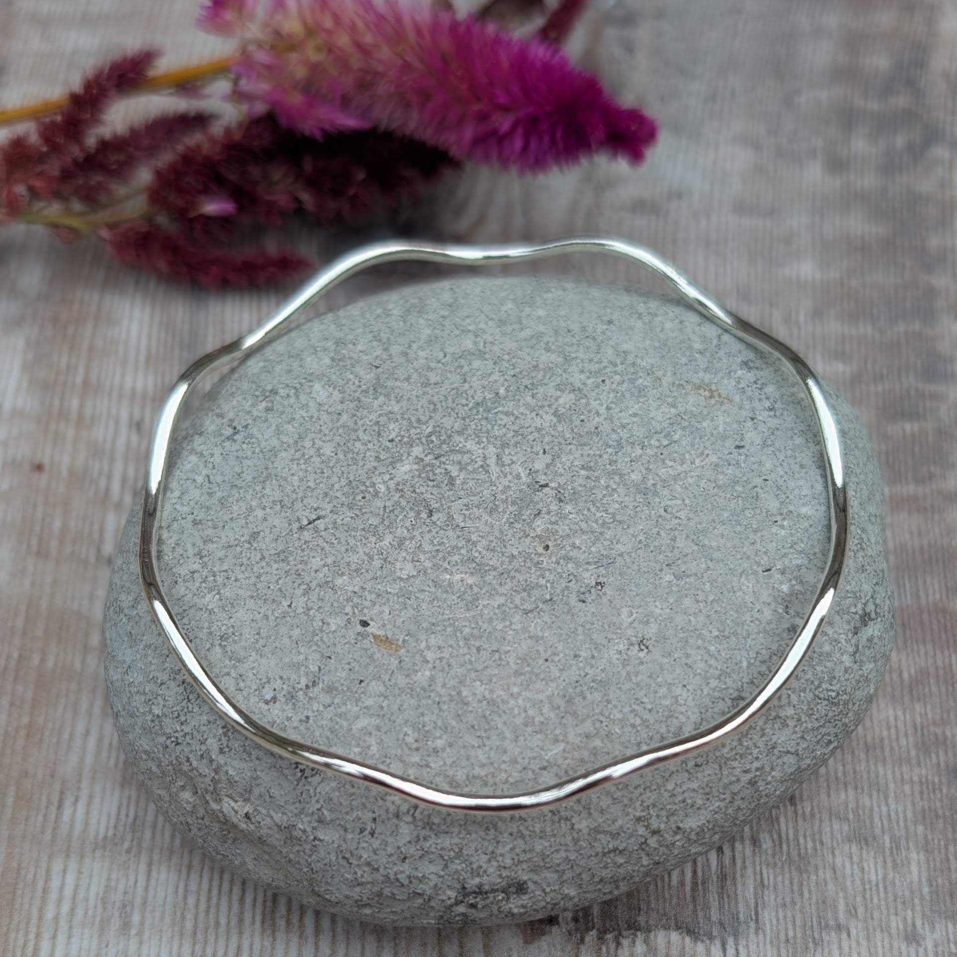 A sterling silver wave bangle resting on a smooth stone, displaying its elegant undulating design and polished surface.