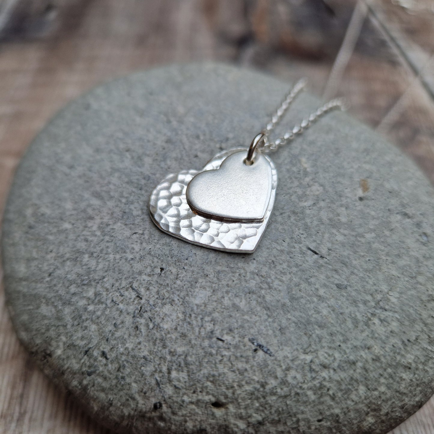 Sterling Silver hammered heart shape hangs from silver earring wire. Smaller heart shape with smooth finish sits on top of the larger hammered heart.