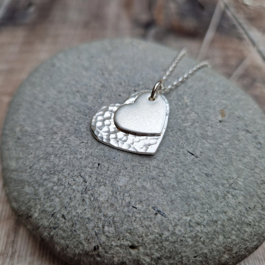 Sterling Silver hammered heart shape hangs from silver earring wire. Smaller heart shape with smooth finish sits on top of the larger hammered heart.