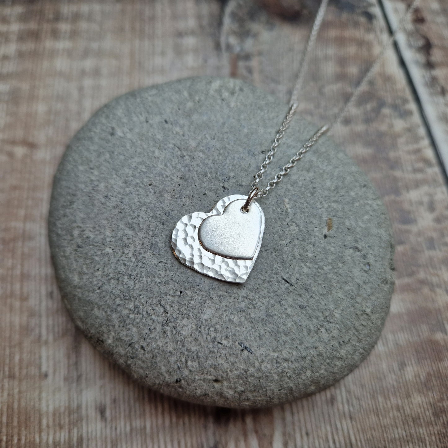 Sterling Silver hammered heart shape hangs from silver earring wire. Smaller heart shape with smooth finish sits on top of the larger hammered heart.