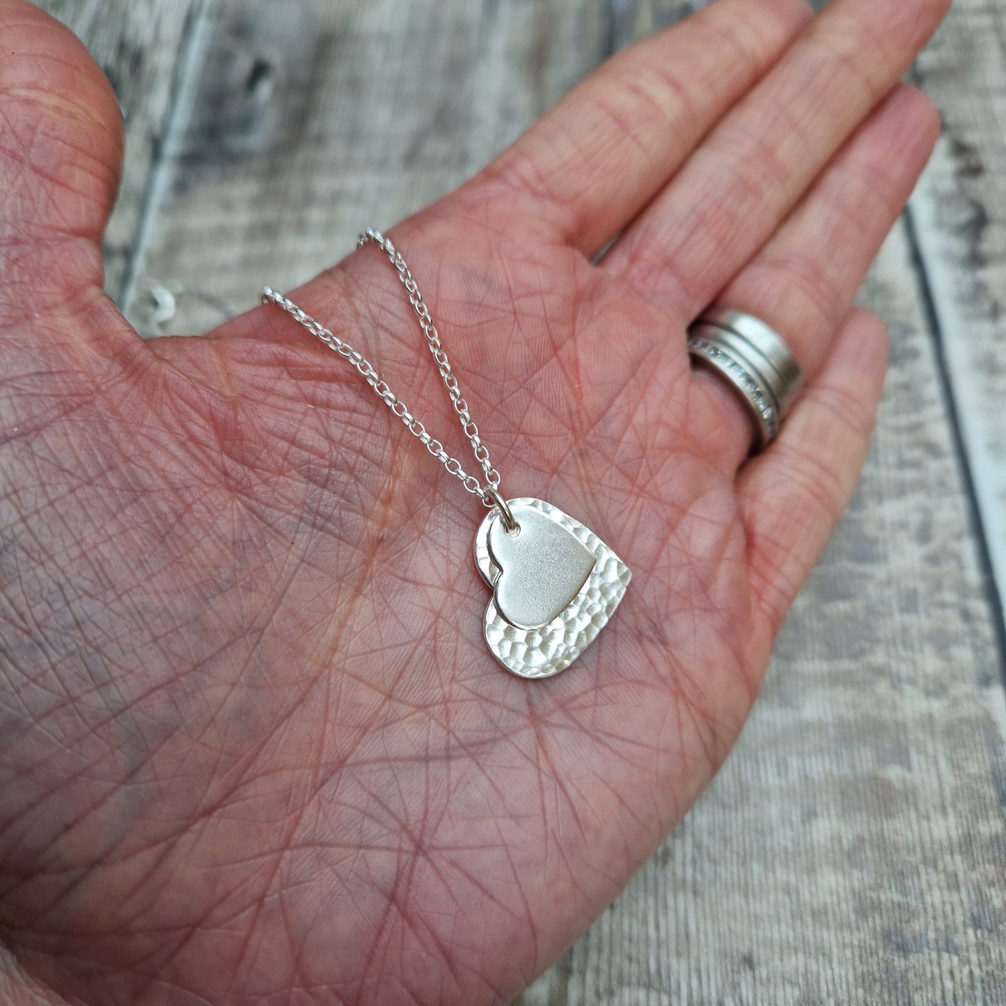 Sterling Silver hammered heart shape hangs from silver earring wire. Smaller heart shape with smooth finish sits on top of the larger hammered heart.