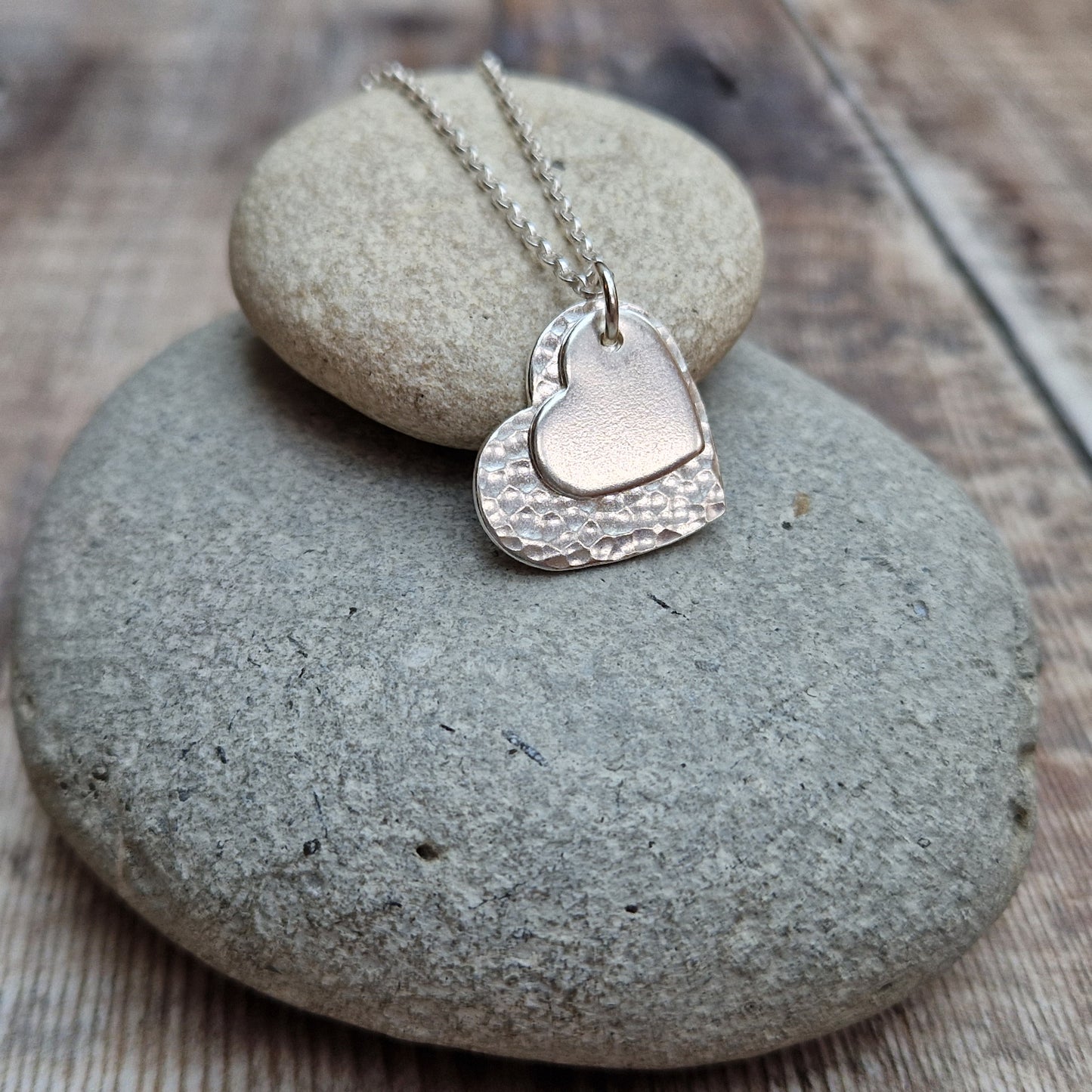 Sterling Silver hammered heart shape hangs from silver earring wire. Smaller heart shape with smooth finish sits on top of the larger hammered heart.