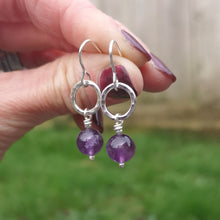 Load image into Gallery viewer, Sterling Silver Amethyst gemstone circle drop earrings. Each earring has one open silver circle with hammered texture, attached to silver earring wire. Attached to the open circle via a silver bar with small twist detail is a sphere of purple Amethyst gemstone with the silver bar passing through the middle and almost visible. Measuring approximately 10mm diameter. Approximately 35mm drop from earlobe including ear hook.
