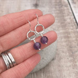Sterling Silver Amethyst gemstone circle drop earrings. Each earring has one open silver circle with hammered texture, attached to silver earring wire. Attached to the open circle via a silver bar with small twist detail is a sphere of purple Amethyst gemstone with the silver bar passing through the middle and almost visible. Measuring approximately 10mm diameter. Approximately 35mm drop from earlobe including ear hook.