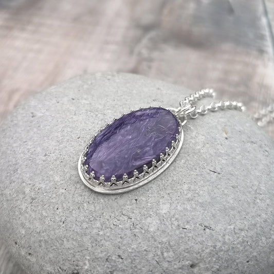 Oval shaped Charoite pendant set in detailed Sterling Silver bezel, attached to chain via two small silver circle links. Charoite stone is dark purple with light purple swirls running through it with polished finish.