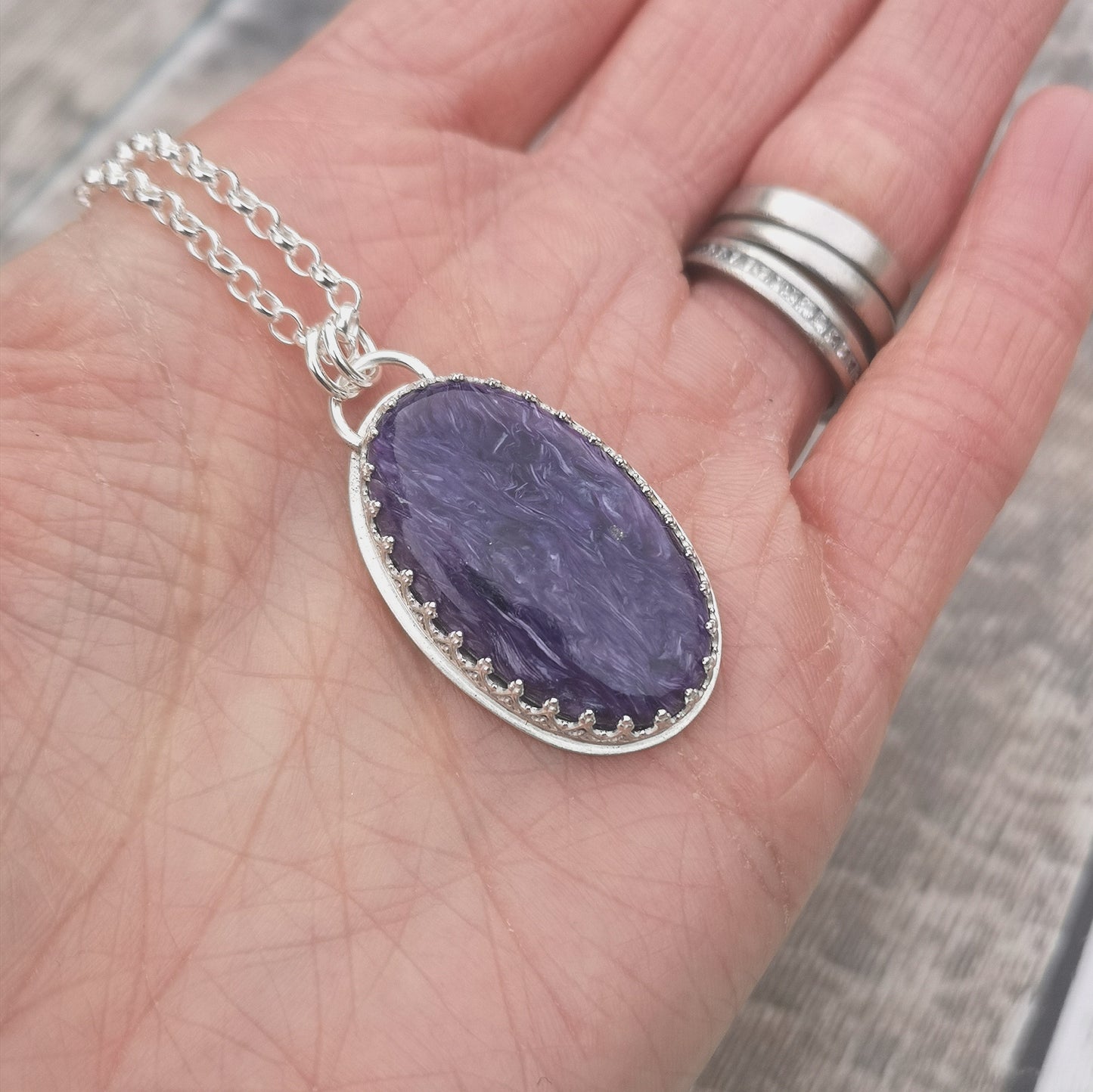 Oval shaped Charoite pendant set in detailed Sterling Silver bezel, attached to chain via two small silver circle links. Charoite stone is dark purple with light purple swirls running through it with polished finish.