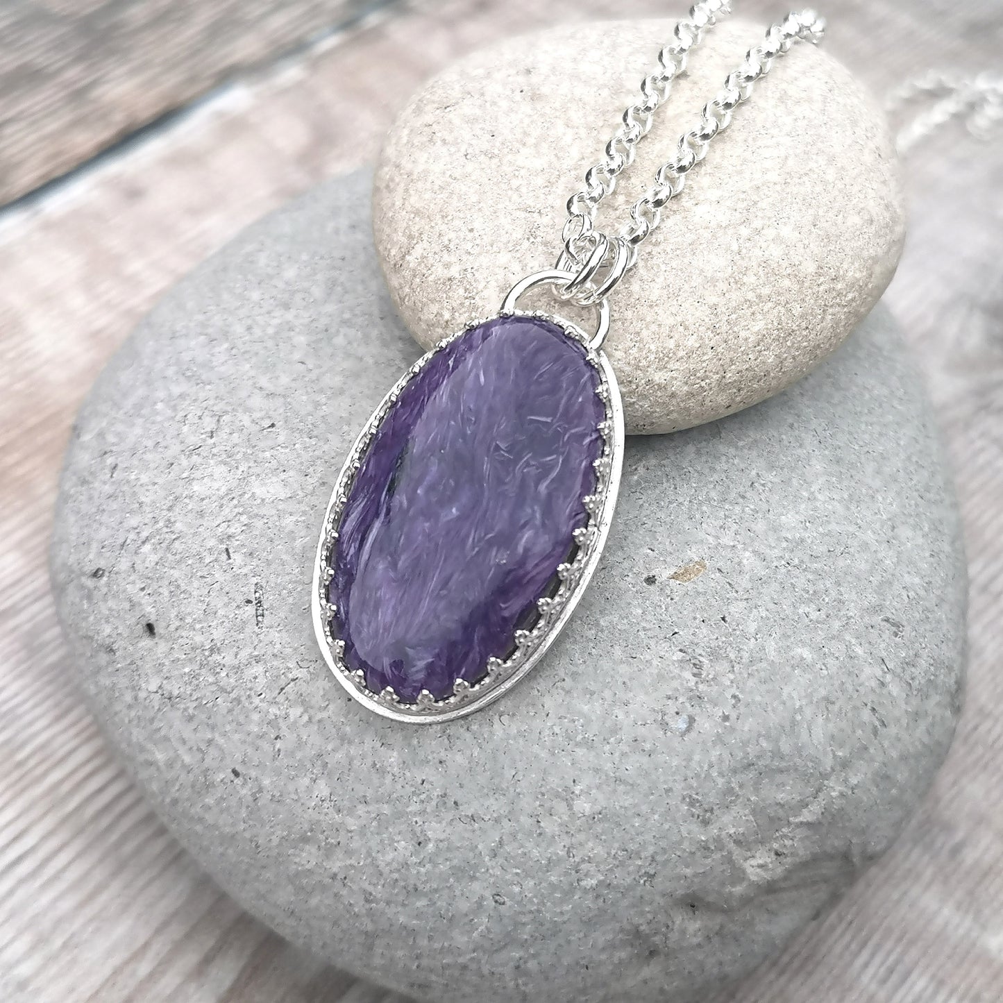 Oval shaped Charoite pendant set in detailed Sterling Silver bezel, attached to chain via two small silver circle links. Charoite stone is dark purple with light purple swirls running through it with polished finish.