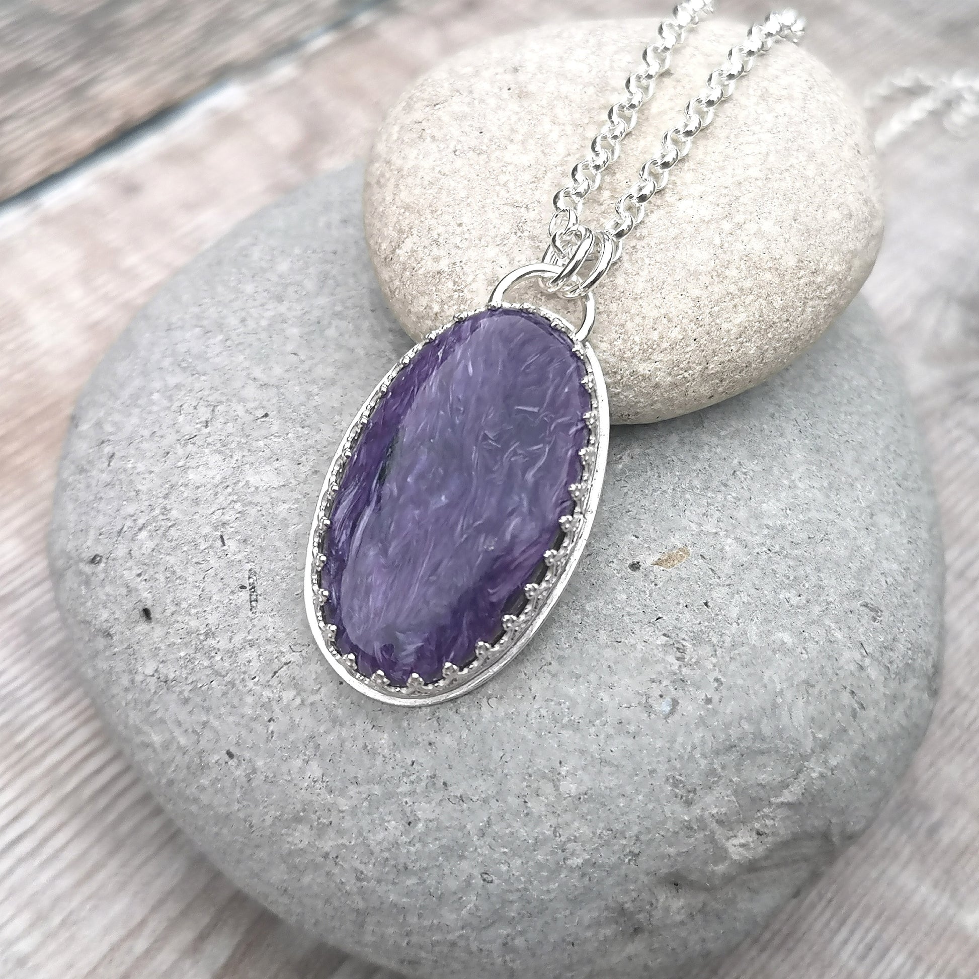 Oval shaped Charoite pendant set in detailed Sterling Silver bezel, attached to chain via two small silver circle links. Charoite stone is dark purple with light purple swirls running through it with polished finish.