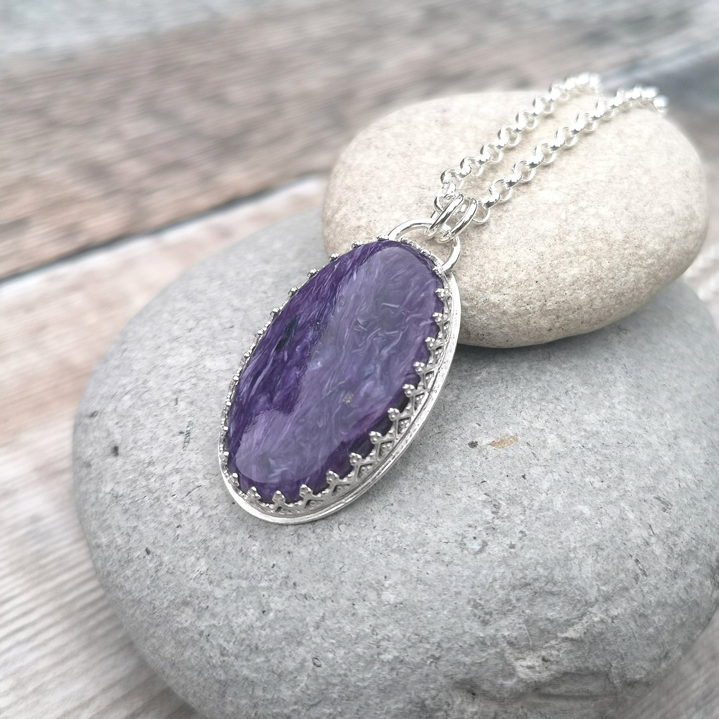 Oval shaped Charoite pendant set in detailed Sterling Silver bezel, attached to chain via two small silver circle links. Charoite stone is dark purple with light purple swirls running through it with polished finish.