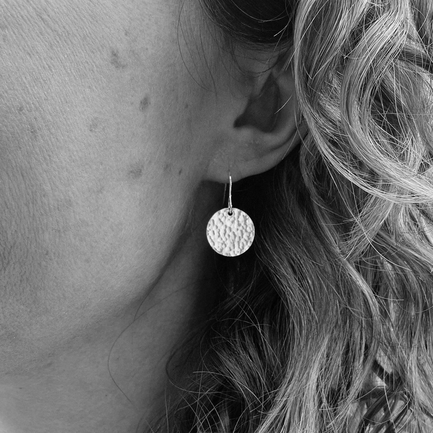 Black and white close-up of a person wearing a pair of silver dangle earrings with hammered disc designs. The textured discs add a touch of shine and texture, hanging elegantly from the ear.