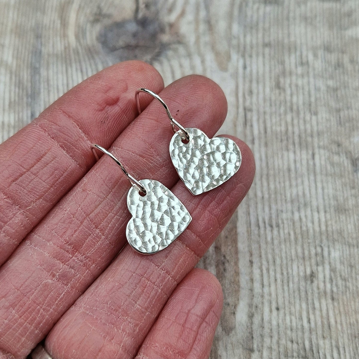 A close-up of two handmade sterling silver heart-shaped earrings with a hammered texture, held between fingers to show their size. The hammered surface gives the hearts a unique, reflective quality, highlighting the craftsmanship of the design.