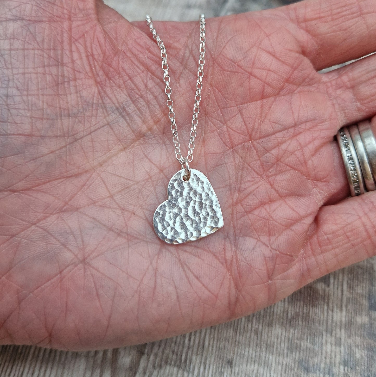 A close-up of a handmade sterling silver heart pendant with a hammered surface, displayed in an open hand to show its size and detail. The pendant is attached to a fine silver chain, reflecting light and showcasing its handcrafted texture.