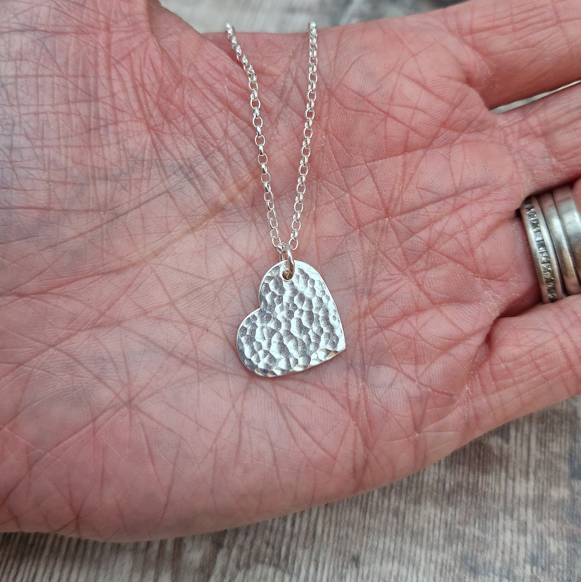 A close-up of a handmade sterling silver heart pendant with a hammered surface, displayed in an open hand to show its size and detail. The pendant is attached to a fine silver chain, reflecting light and showcasing its handcrafted texture.