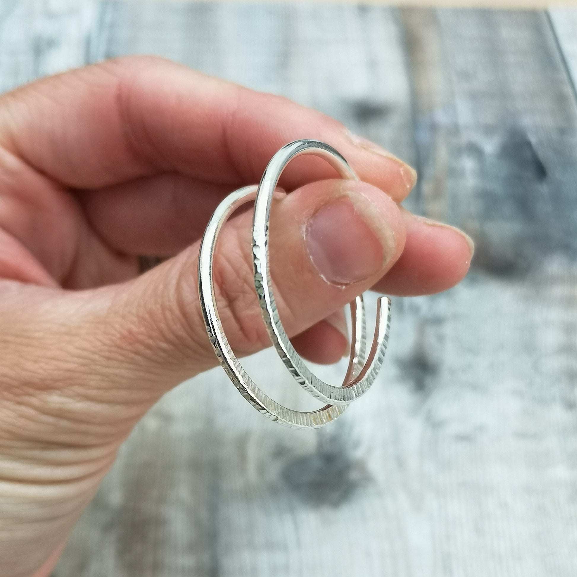 Hand holding large handmade sterling silver hoop earrings, showing the hammered texture and size of the hoops against a rustic wooden background.