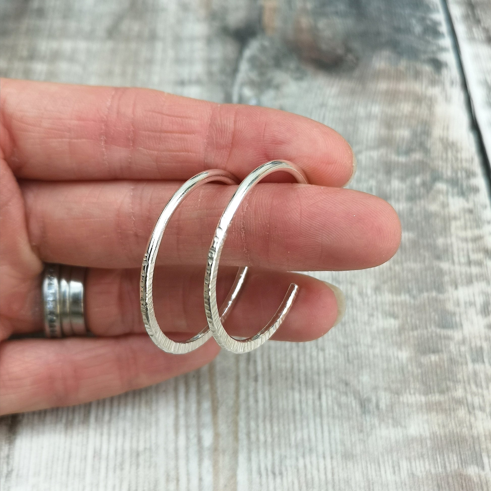 Hand holding large handmade sterling silver hoop earrings, showing the hammered texture and size of the hoops against a rustic wooden background.