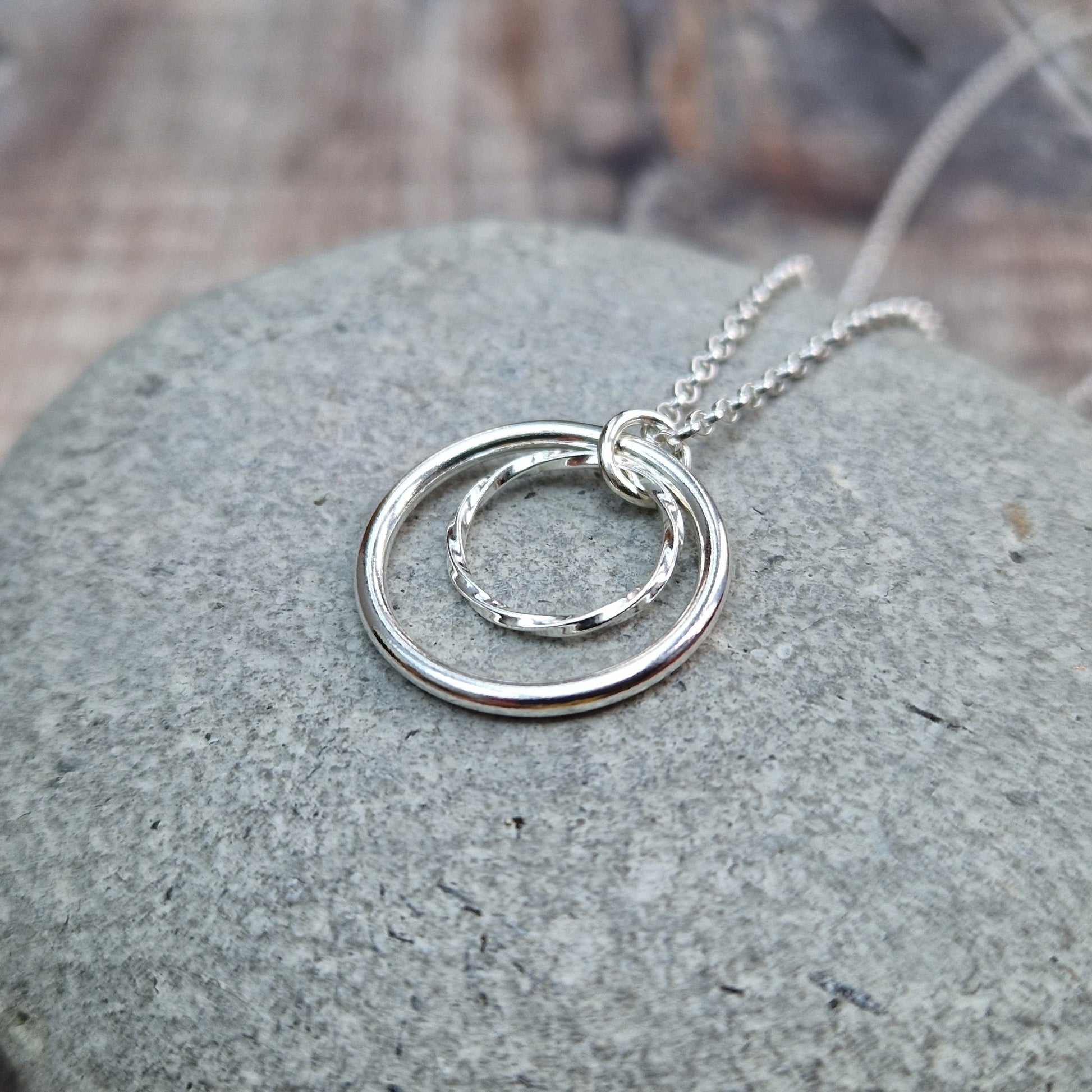 Pendant is a smooth silver open circle and a smaller textured open circle suspended from a silver chain via a small open circle link. The textured circle sits inside the smooth circle.
