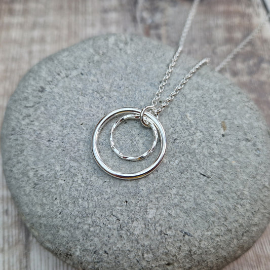 Pendant is a smooth silver open circle and a smaller textured open circle suspended from a silver chain via a small open circle link. The textured circle sits inside the smooth circle.