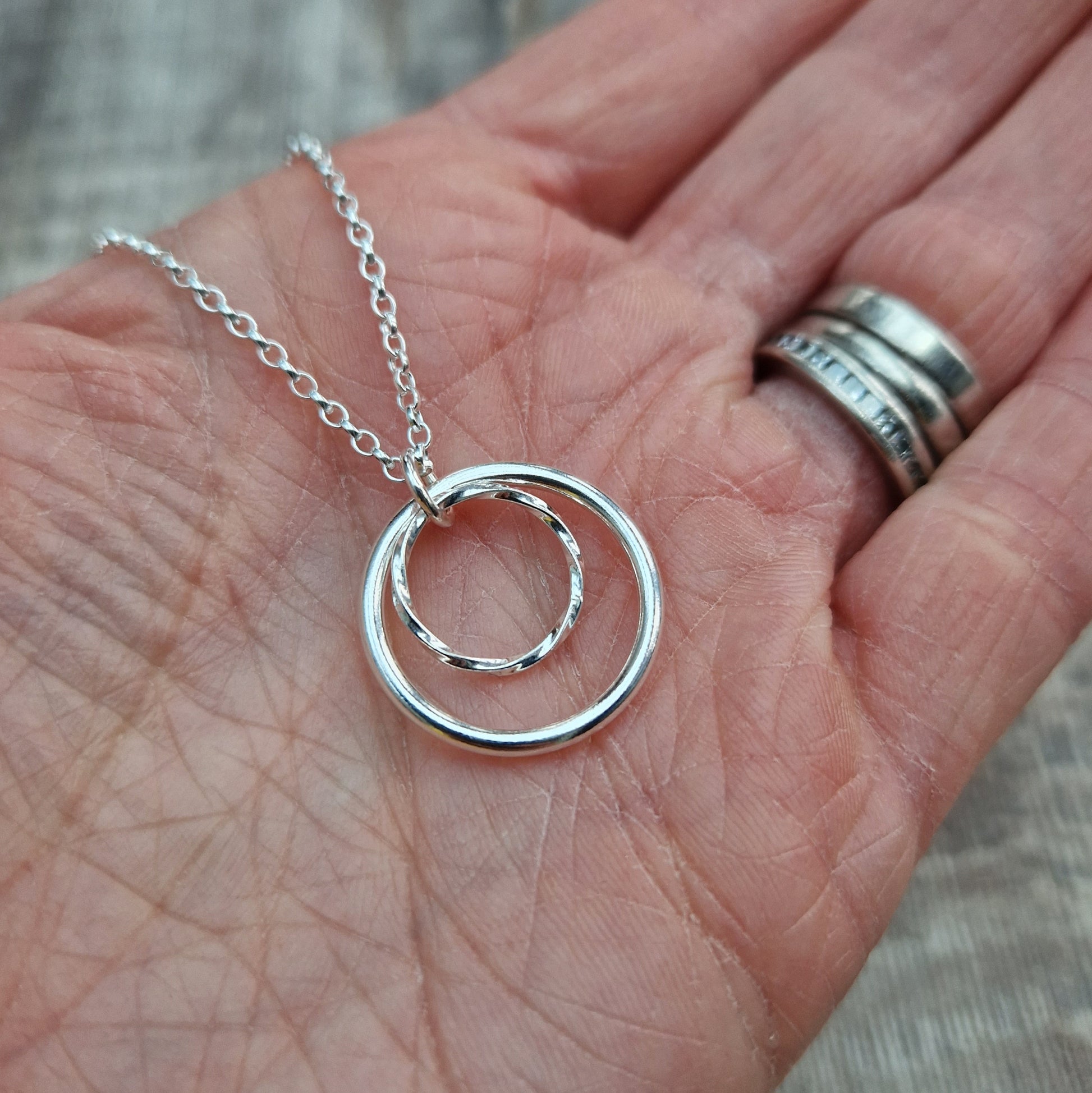 Pendant is a smooth silver open circle and a smaller textured open circle suspended from a silver chain via a small open circle link. The textured circle sits inside the smooth circle.