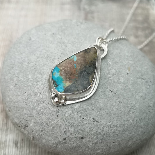 Pendant is a pear cut turquoise gemstone set in Sterling Silver bezel with small three silver sphere shapes set at the bottom of the bezel. Suspended from a silver chain via D-Link. The turquoise stone is blue with large plumes of black and brown.