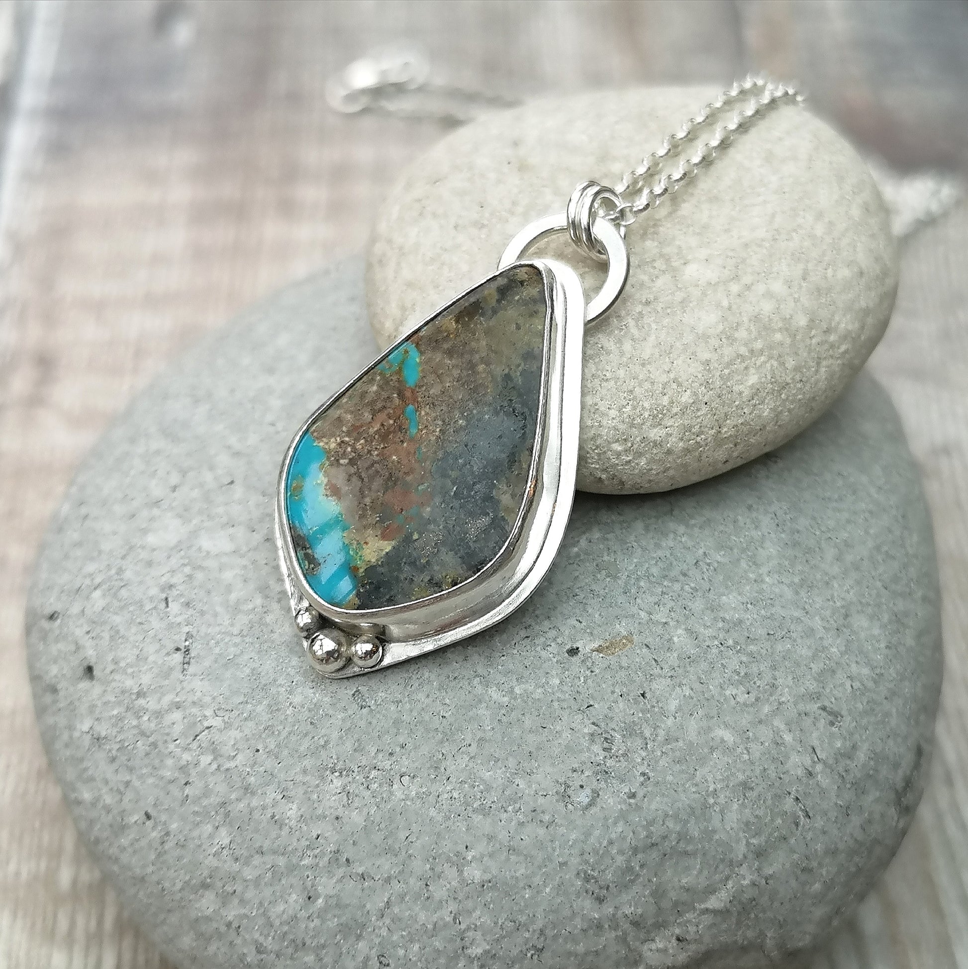 Pendant is a pear cut turquoise gemstone set in Sterling Silver bezel with small three silver sphere shapes set at the bottom of the bezel. Suspended from a silver chain via D-Link. The turquoise stone is blue with large plumes of black and brown.