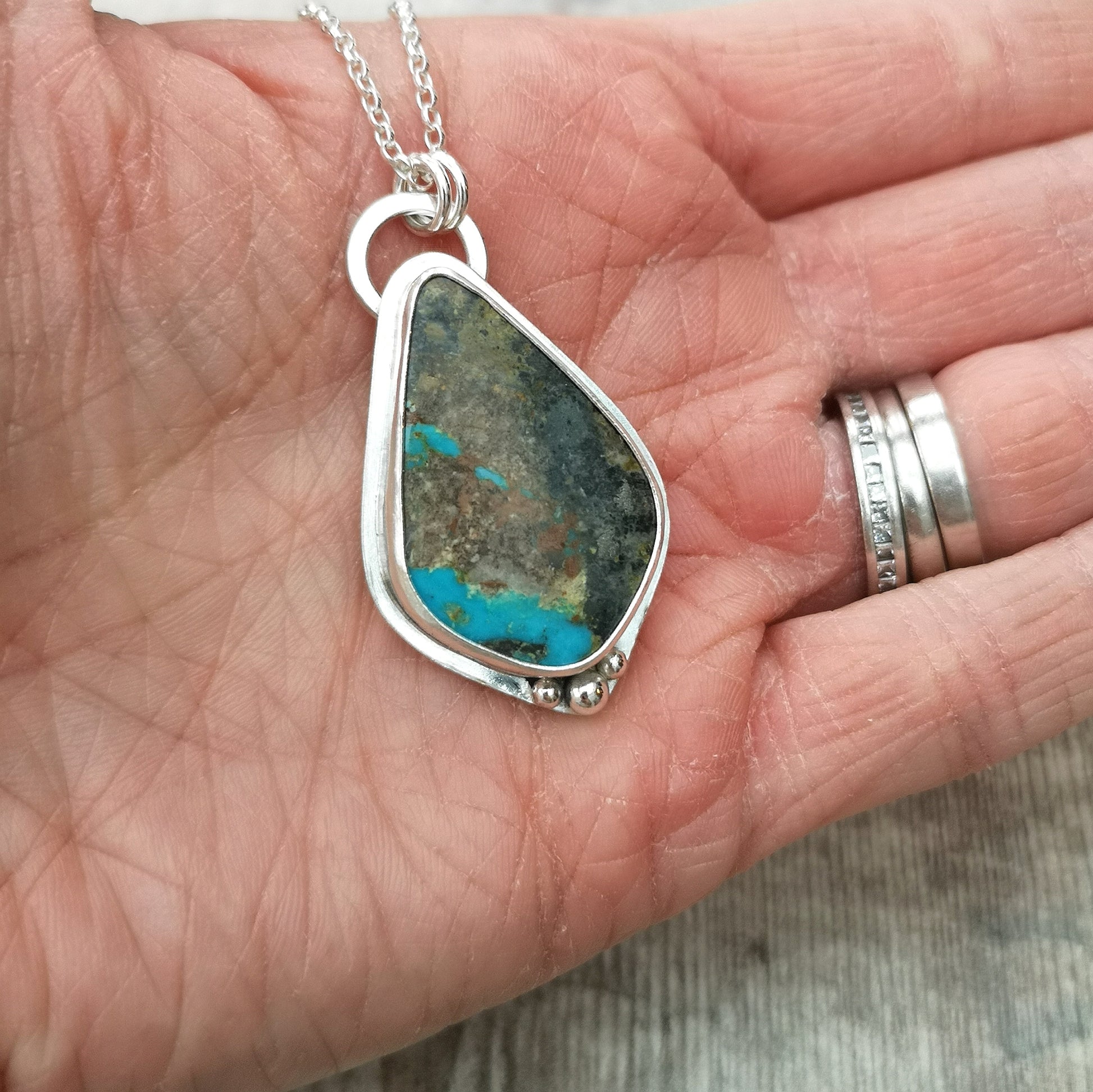 Pendant is a pear cut turquoise gemstone set in Sterling Silver bezel with small three silver sphere shapes set at the bottom of the bezel. Suspended from a silver chain via D-Link. The turquoise stone is blue with large plumes of black and brown.