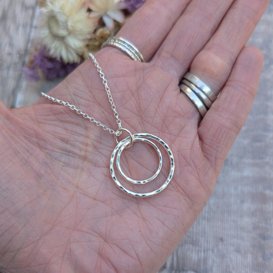 Silver double circle necklace being shown held in the palm of a hand with dried flowers in the background. 