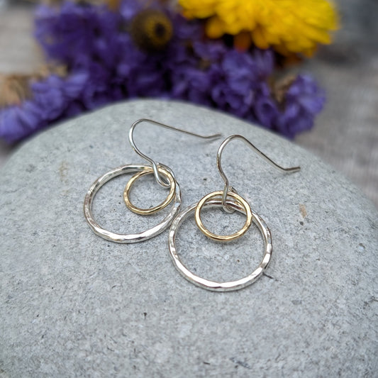 A pair of handmade earrings featuring two circles, one in silver and the other in gold, displayed on a smooth stone surface. The background includes a soft blur of purple and yellow flowers, enhancing the delicate craftsmanship of the earrings.