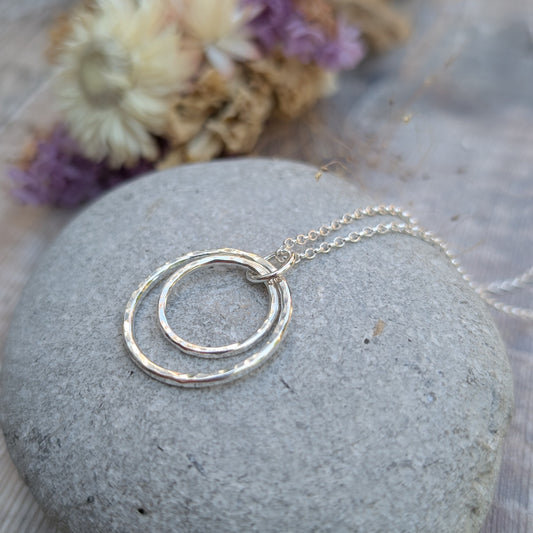 A handmade silver necklace with two interlinked hammered circles resting on a smooth grey stone. The necklace features a delicate silver chain and is styled with soft-focus dried flowers in the background.