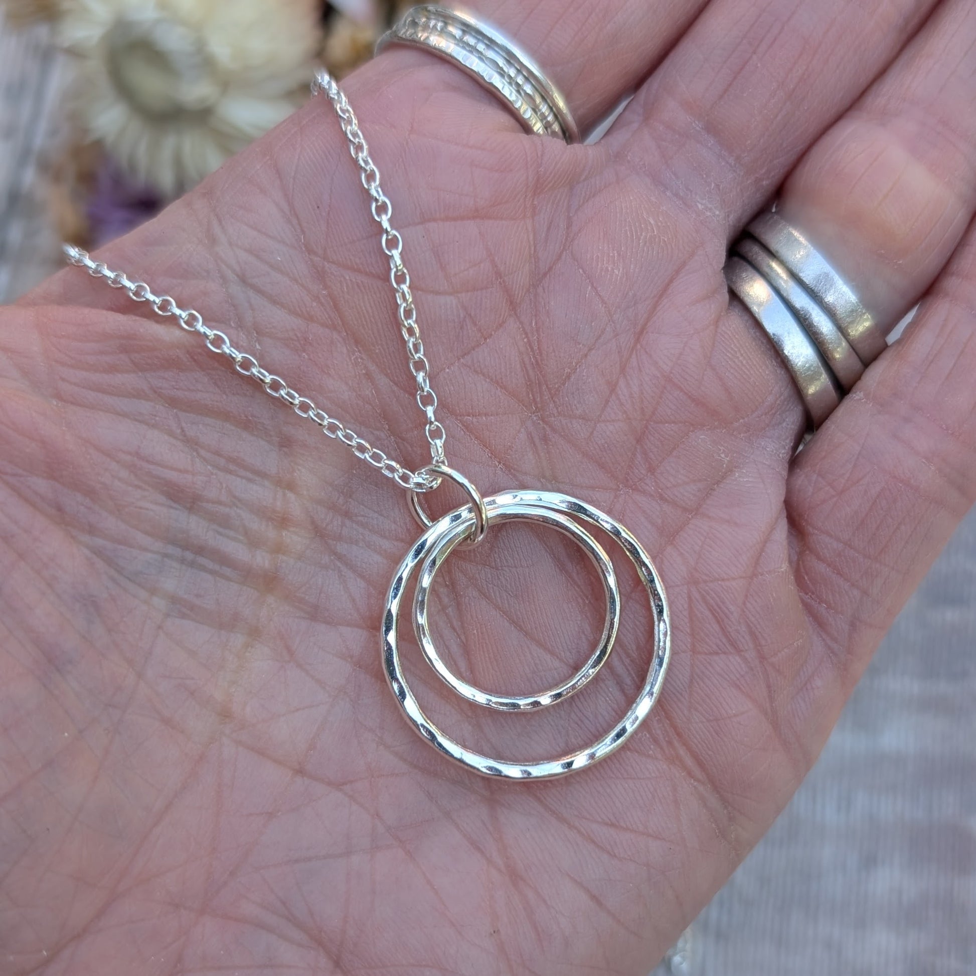 A close-up of a hand holding a handmade silver necklace with two hammered interlinked circles. The silver chain is looped through the top circle, and the hand is adorned with multiple silver rings.