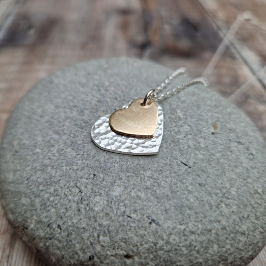 A layered heart necklace featuring a hammered silver heart and a smaller matte gold heart, displayed on a smooth stone surface.