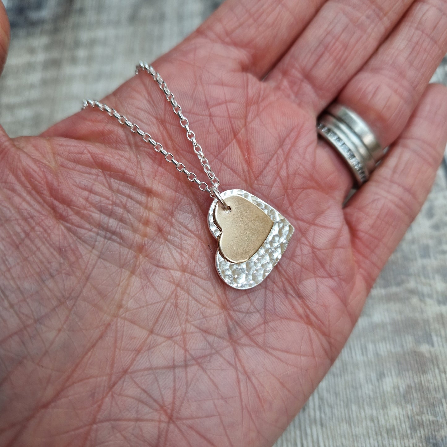 A hand holding the layered heart necklace with a silver and gold heart pendant, highlighting the contrasting textures and finishes.