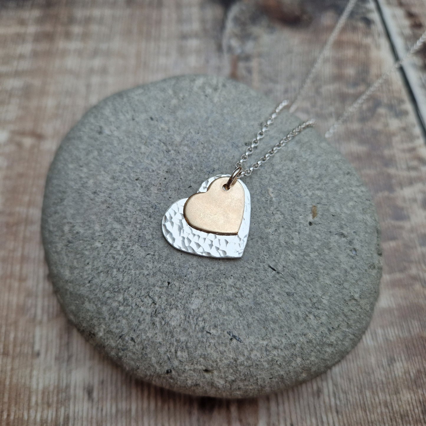 A layered heart necklace featuring a hammered silver heart and a smaller matte gold heart, displayed on a smooth stone surface.