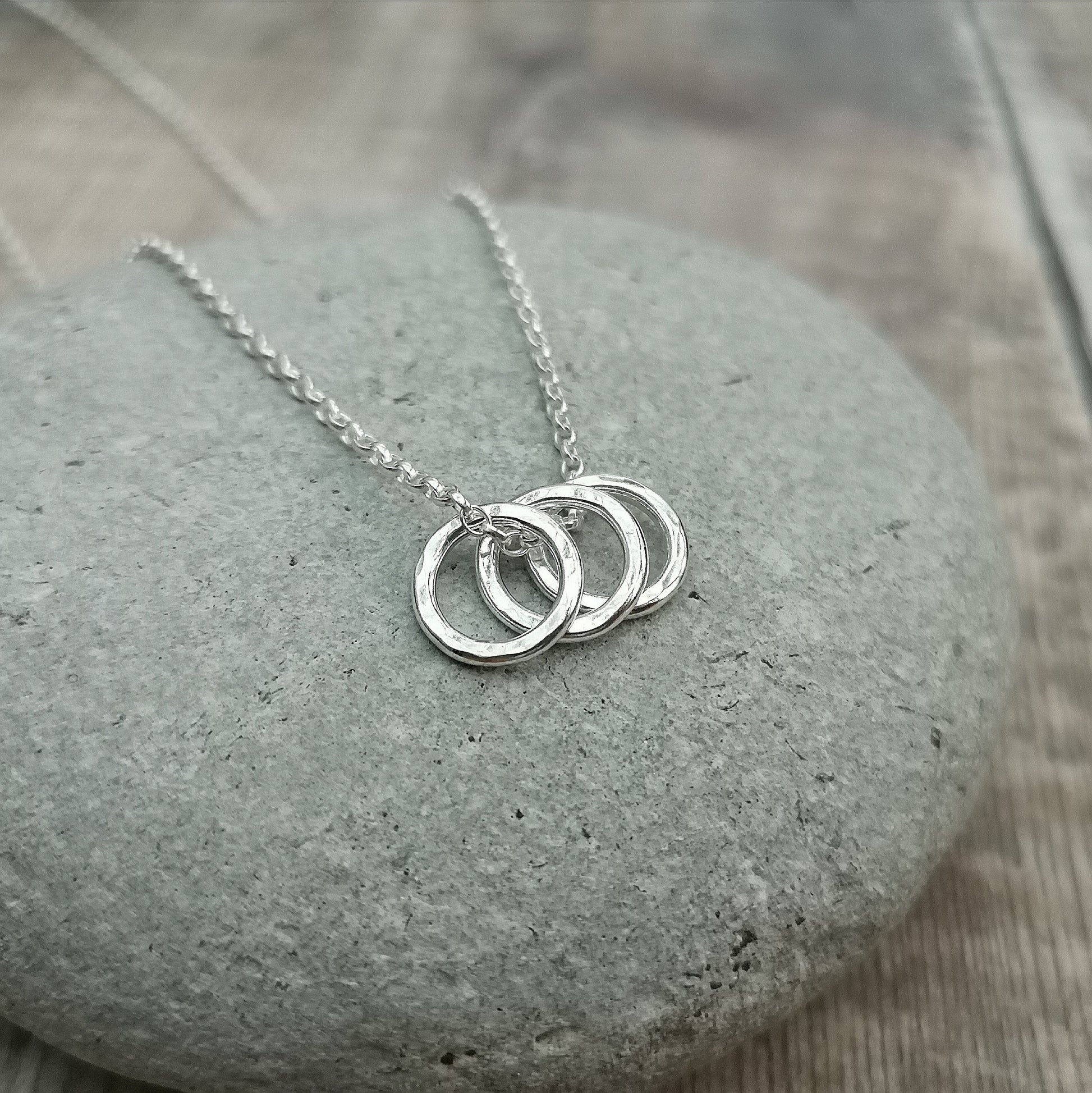 A close-up of a sterling silver necklace with three small hammered rings, resting on a smooth grey stone. The minimalist design and subtle texture of the rings are highlighted by the polished silver finish.