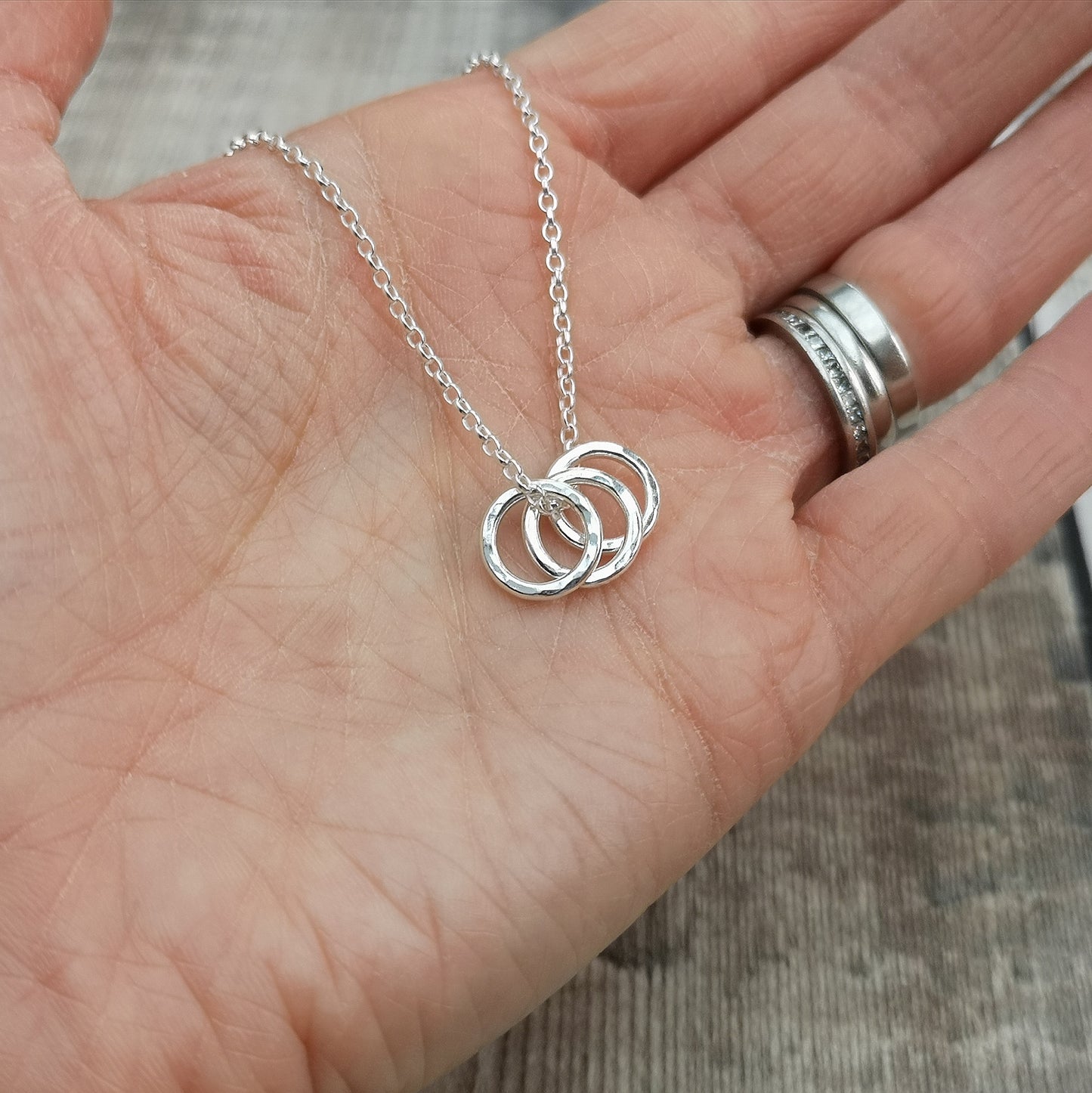 A hand holding a sterling silver necklace featuring three hammered rings, showcasing the small and delicate design of the necklace. The rings dangle lightly from a fine silver chain, creating a simple yet elegant look.