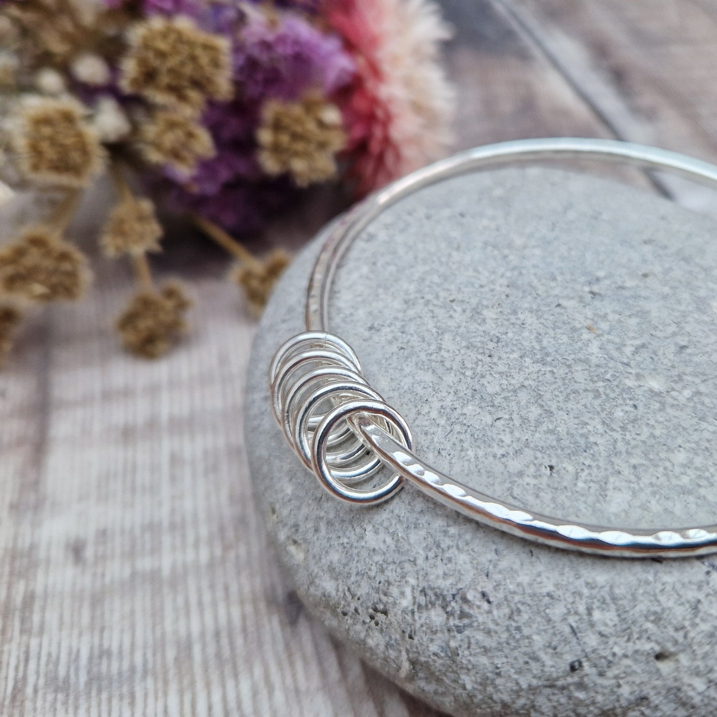 A handmade sterling silver bangle with five small silver rings resting on its hammered surface. The bangle is placed on a smooth stone, with a soft background of dried flowers in purple, pink, and white hues.