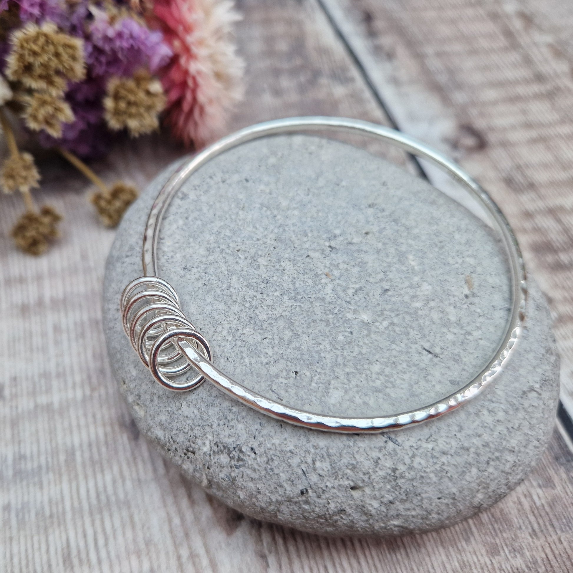 A handmade sterling silver bangle with five small silver rings resting on its hammered surface. The bangle is placed on a smooth stone, with a soft background of dried flowers in purple, pink, and white hues.