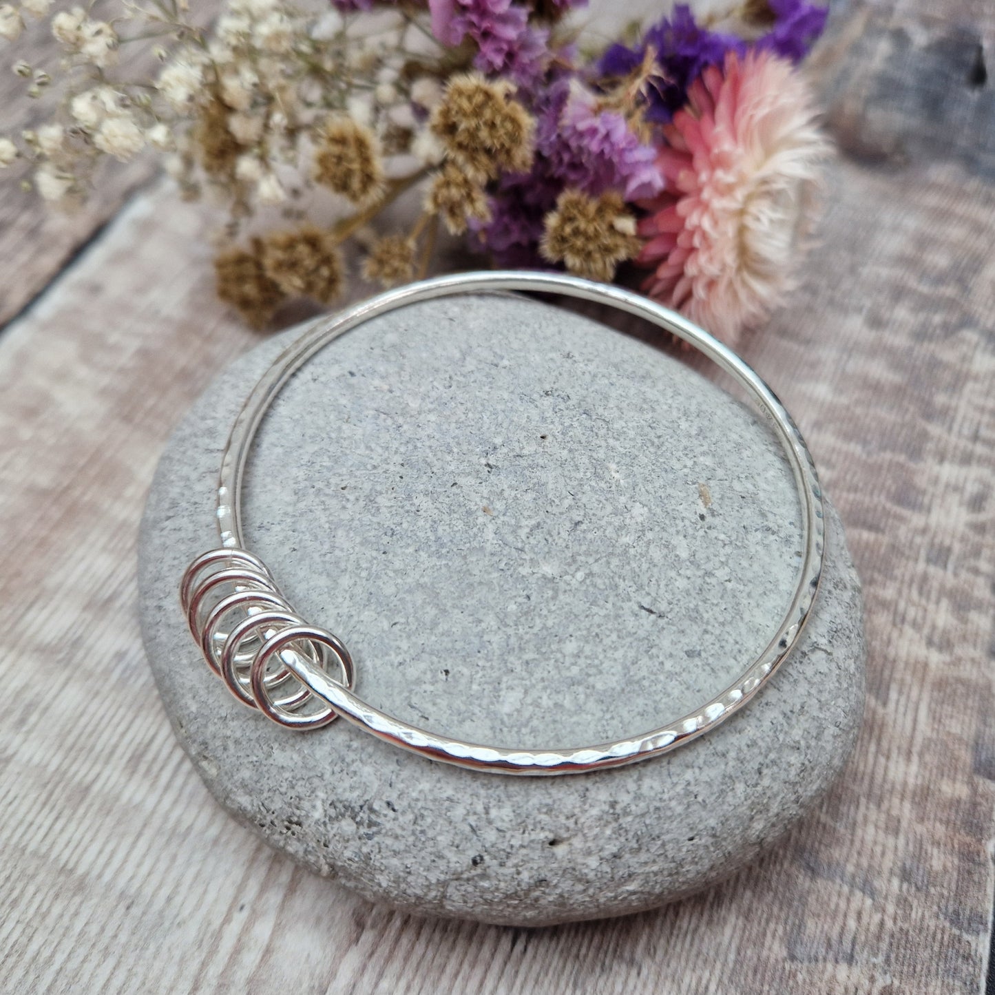 A handmade sterling silver bangle with five small silver rings resting on its hammered surface. The bangle is placed on a smooth stone, with a soft background of dried flowers in purple, pink, and white hues.