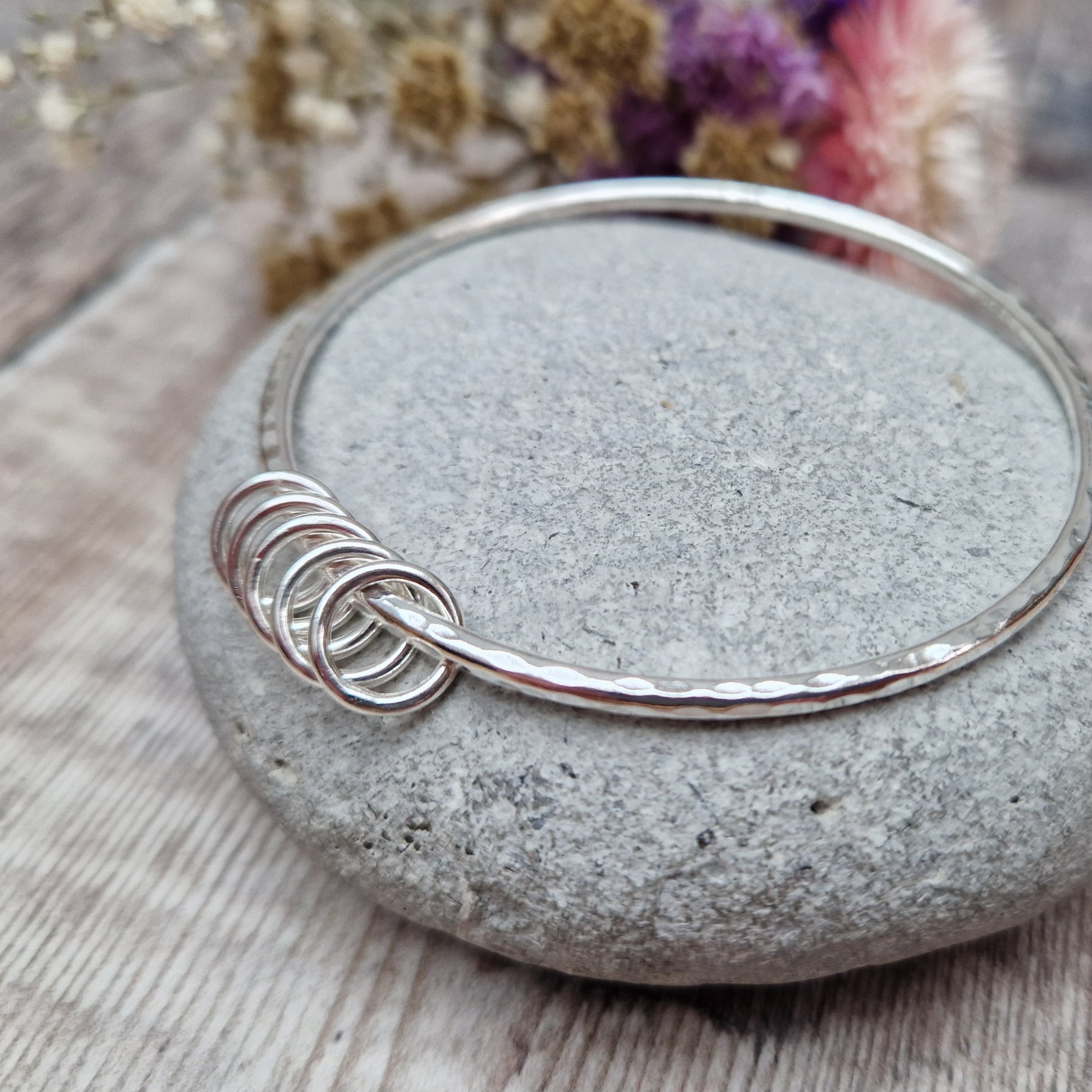 A handmade sterling silver bangle with five small silver rings resting on its hammered surface. The bangle is placed on a smooth stone, with a soft background of dried flowers in purple, pink, and white hues.