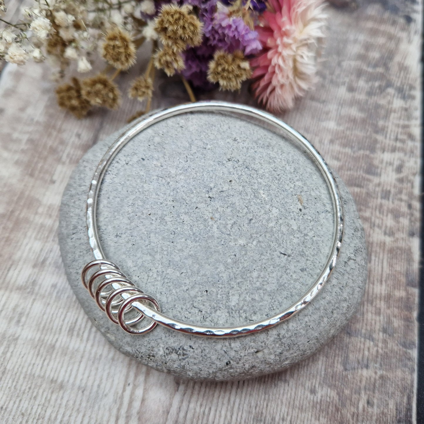 A handmade sterling silver bangle with five small silver rings resting on its hammered surface. The bangle is placed on a smooth stone, with a soft background of dried flowers in purple, pink, and white hues.