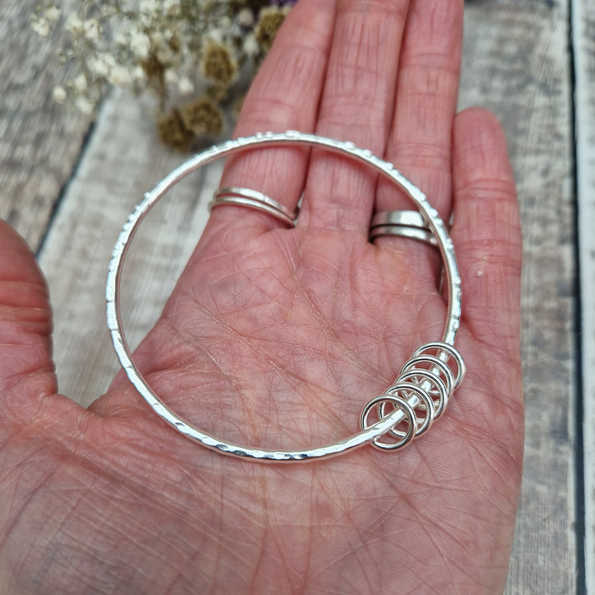 A handmade sterling silver bangle with five silver rings held in an open hand, showing the size and scale of the bangle. The hammered texture of the bangle glimmers against the natural light, while the hand holding it has two silver rings on the fingers.