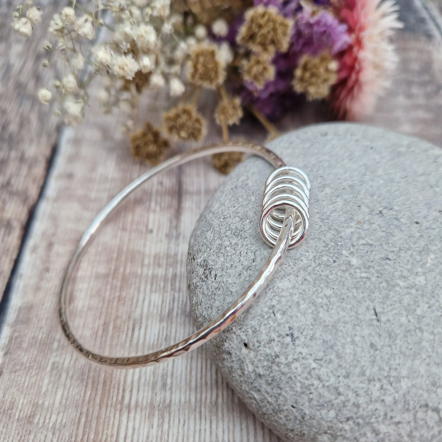 A handmade sterling silver bangle with five small silver rings resting on its hammered surface. The bangle is placed on a smooth stone, with a soft background of dried flowers in purple, pink, and white hues.