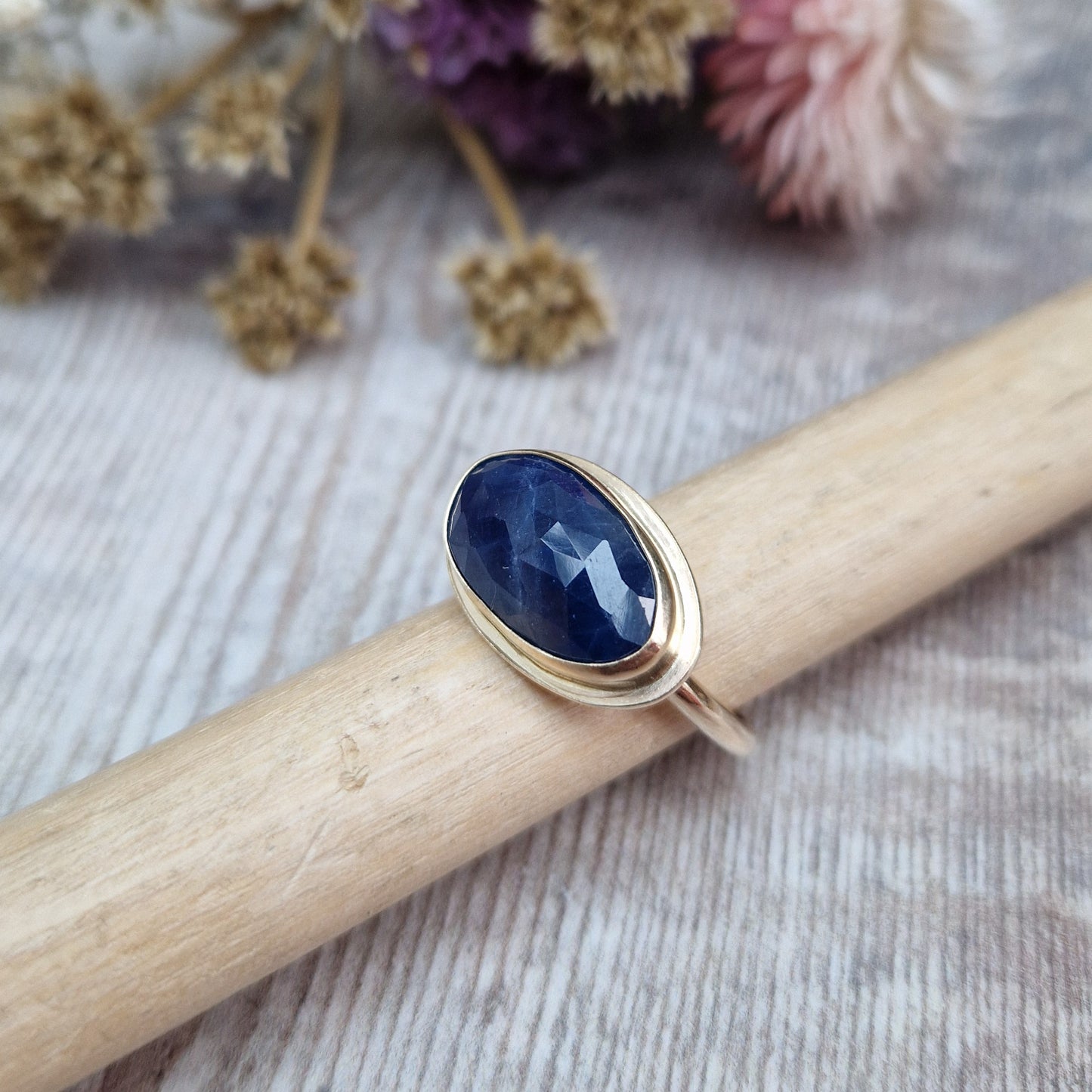 9ct gold ring with dark blue colour sapphire gemstone, oval shaped. Set in raised gold surround. Gemstone set lengthways across ring. Slight texture to stone, like diamond cut.