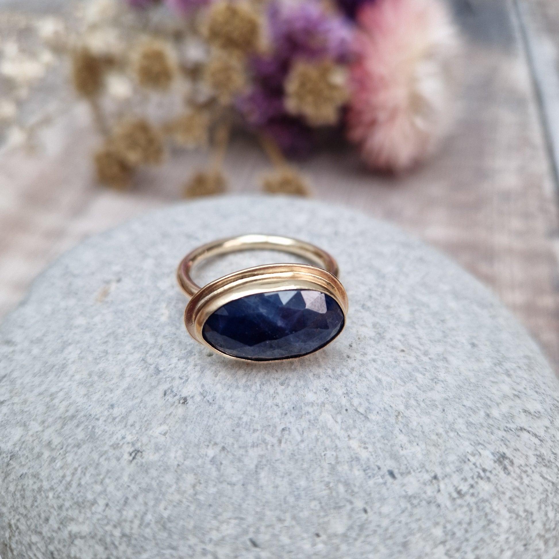 9ct gold ring with dark blue colour sapphire gemstone, oval shaped. Set in raised gold surround. Gemstone set lengthways across ring. Slight texture to stone, like diamond cut.