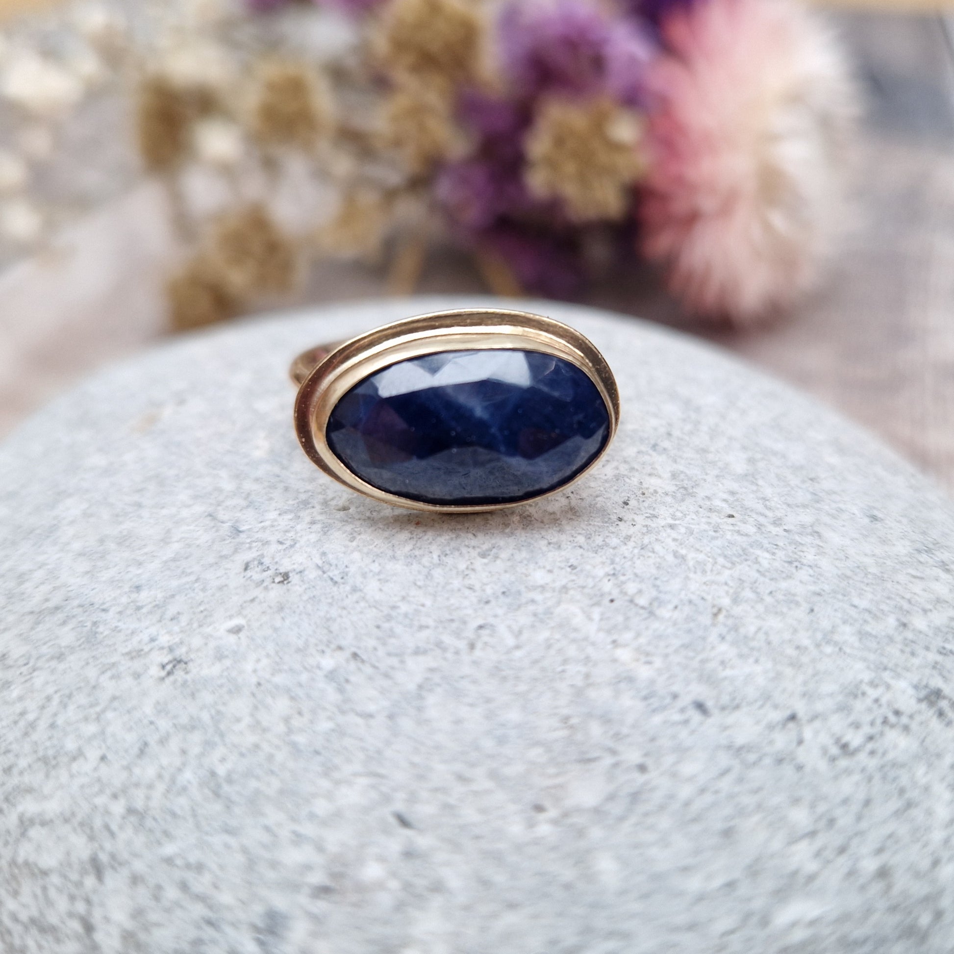 9ct gold ring with dark blue colour sapphire gemstone, oval shaped. Set in raised gold surround. Gemstone set lengthways across ring. Slight texture to stone, like diamond cut.