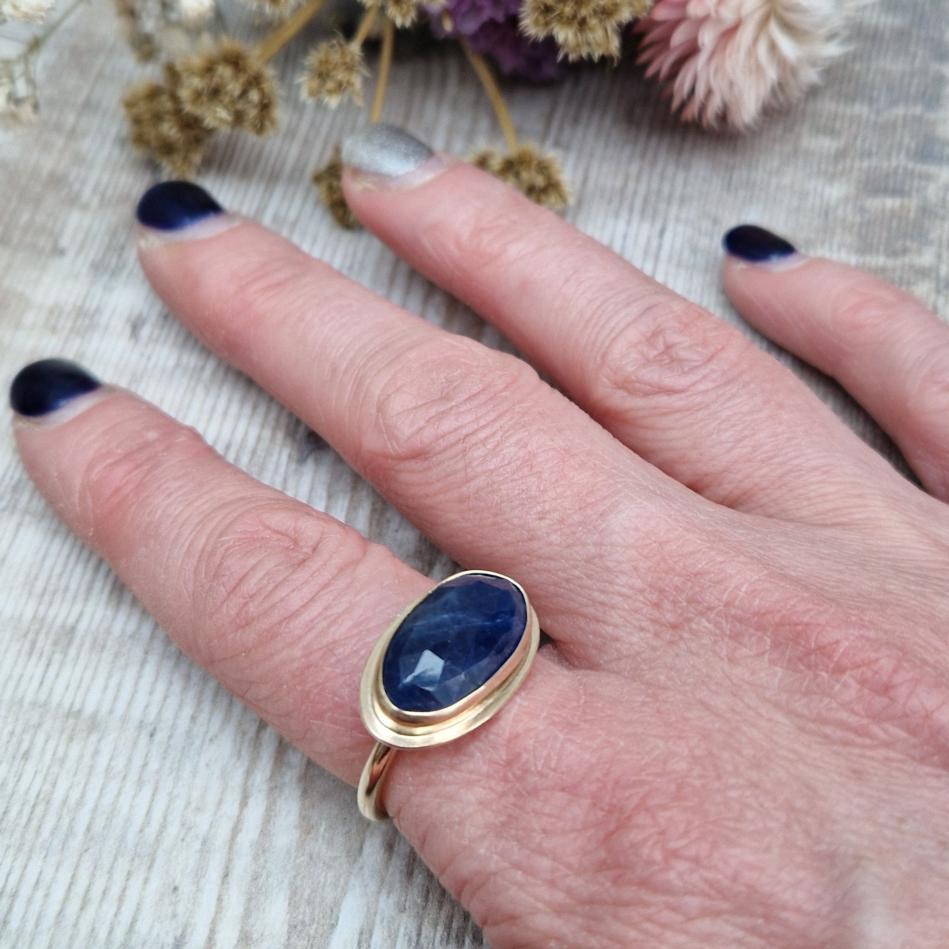 9ct gold ring with dark blue colour sapphire gemstone, oval shaped. Set in raised gold surround. Gemstone set lengthways across ring. Slight texture to stone, like diamond cut.