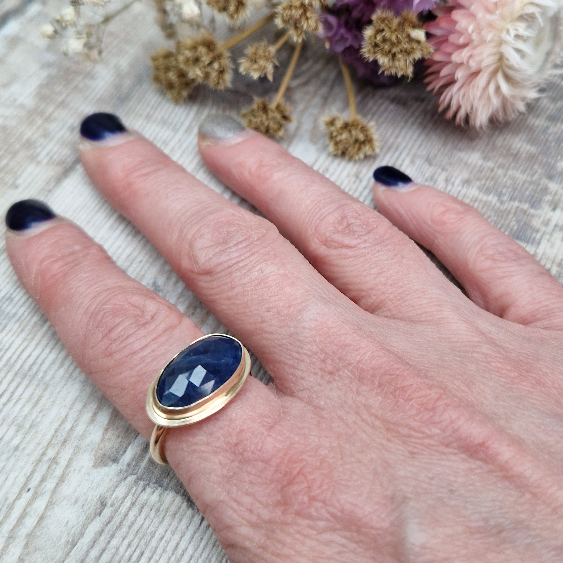 9ct gold ring with dark blue colour sapphire gemstone, oval shaped. Set in raised gold surround. Gemstone set lengthways across ring. Slight texture to stone, like diamond cut.