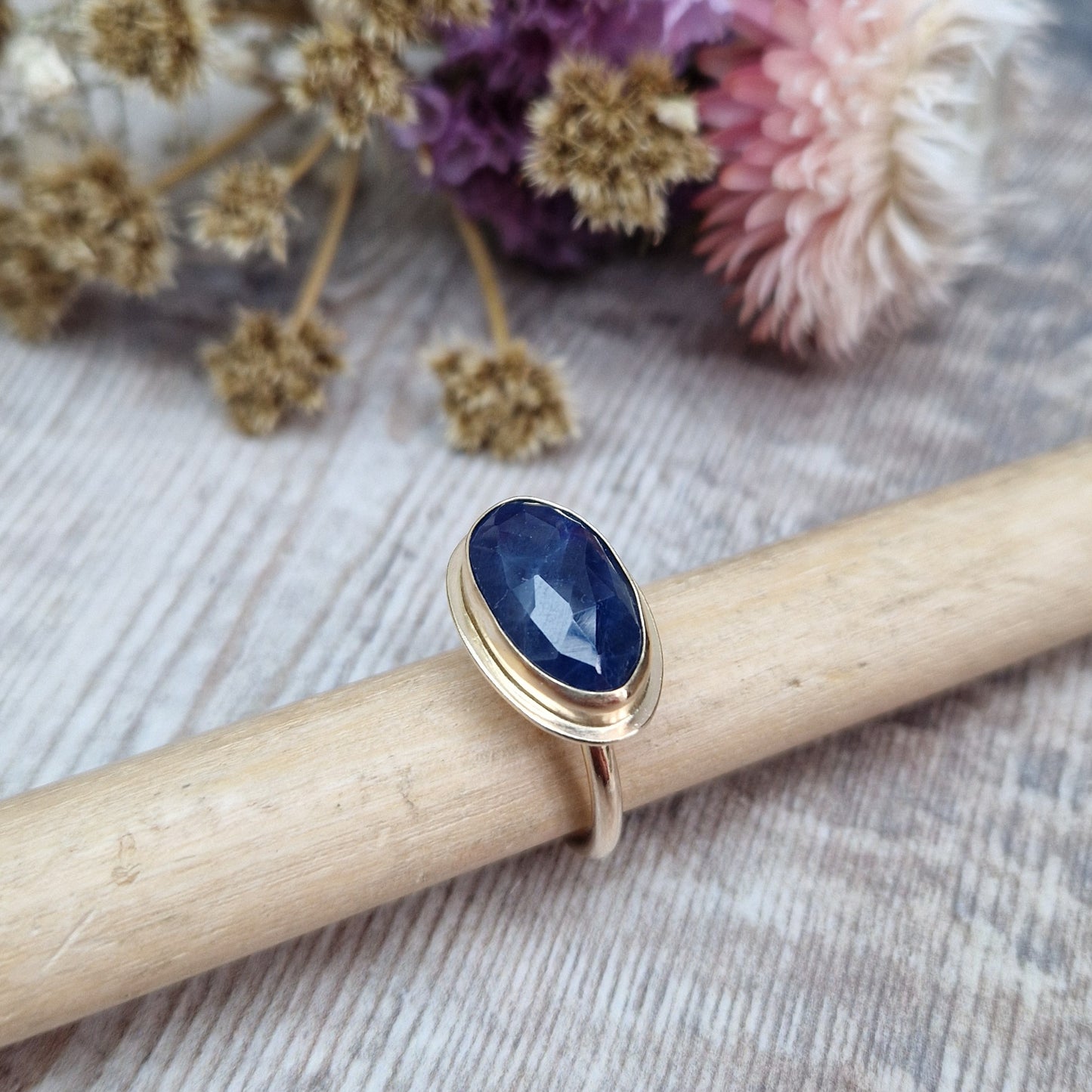 9ct gold ring with dark blue colour sapphire gemstone, oval shaped. Set in raised gold surround. Gemstone set lengthways across ring. Slight texture to stone, like diamond cut.