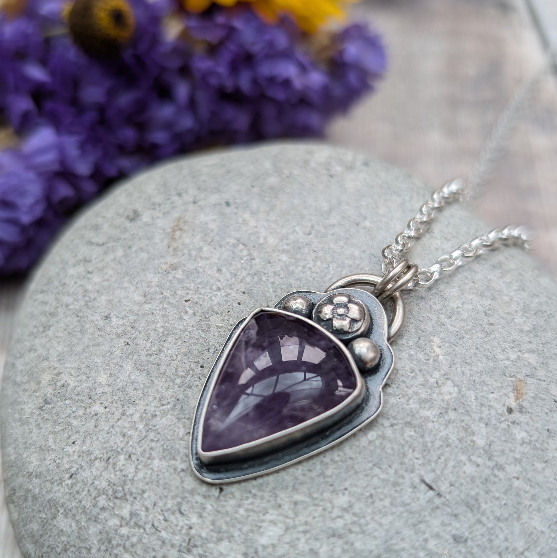 An oxidised silver necklace with a teardrop-shaped amethyst stone pendant. The pendant features a flower and bead design above the dark purple stone on the bezel setting.