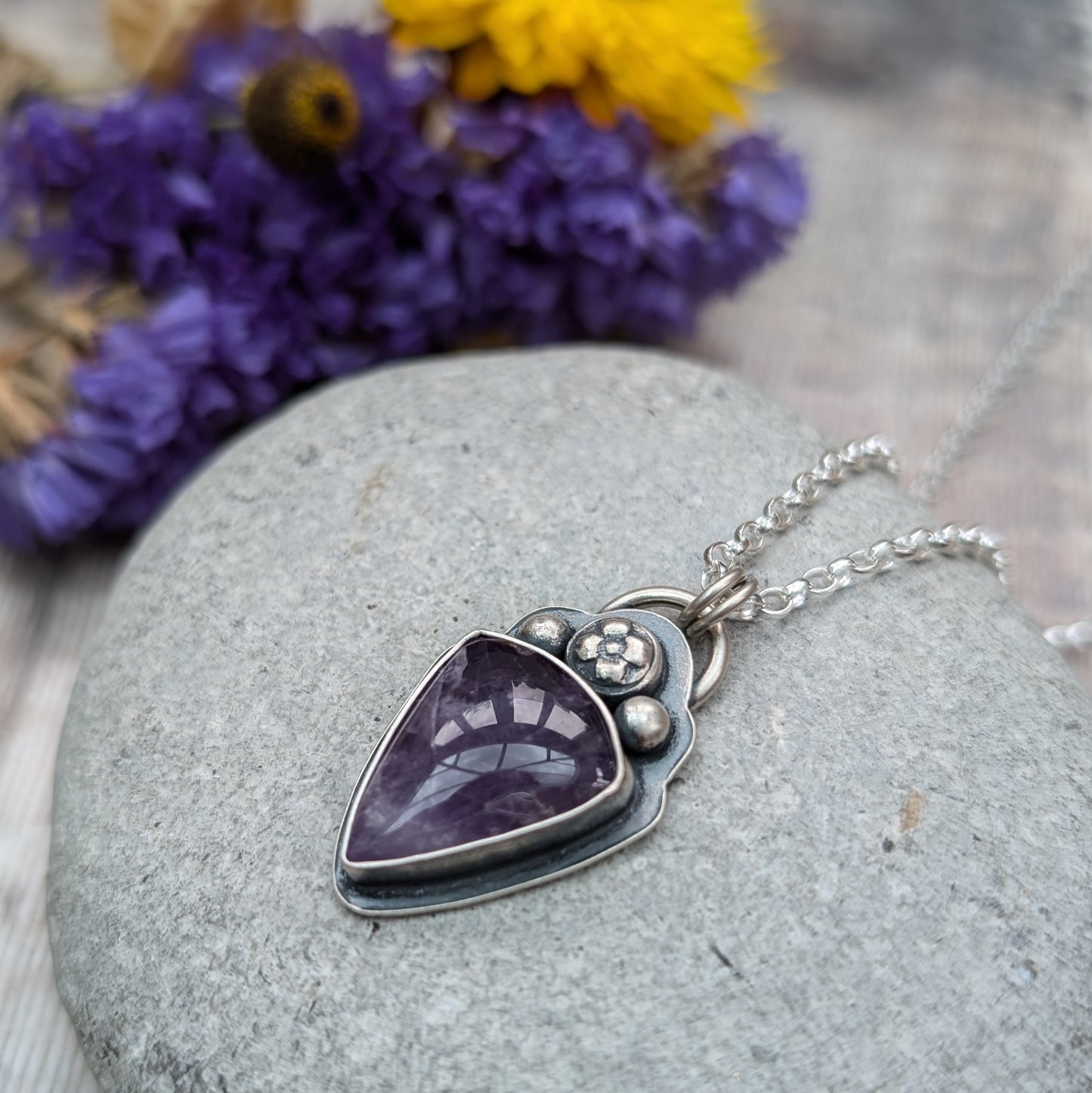 An oxidised silver necklace with a teardrop-shaped amethyst stone pendant. The pendant features a flower and bead design above the dark purple stone on the bezel setting.