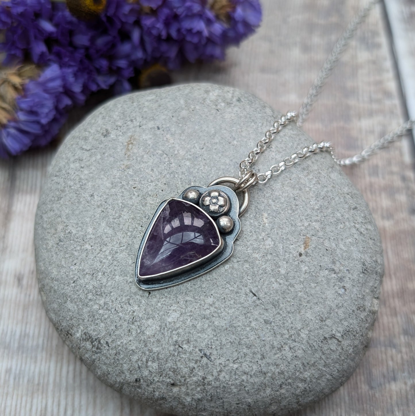 An oxidised silver necklace with a teardrop-shaped amethyst stone pendant. The pendant features a flower and bead design above the dark purple stone on the bezel setting.